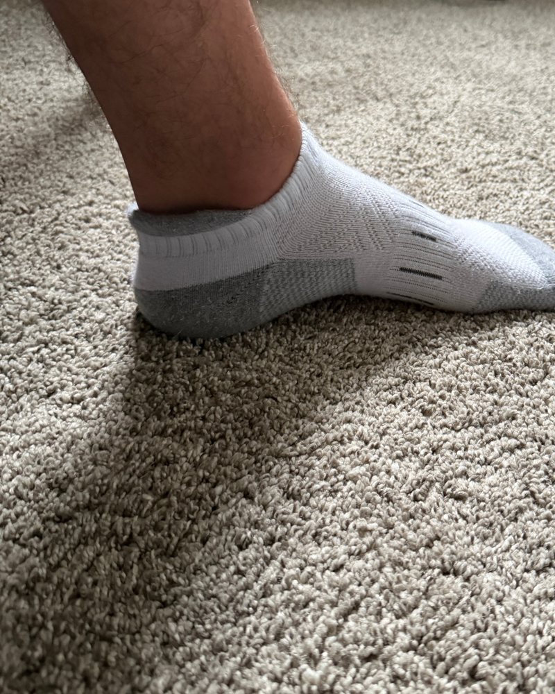 Phone photo of a white ankle sock worn on a foot, standing on carpet.